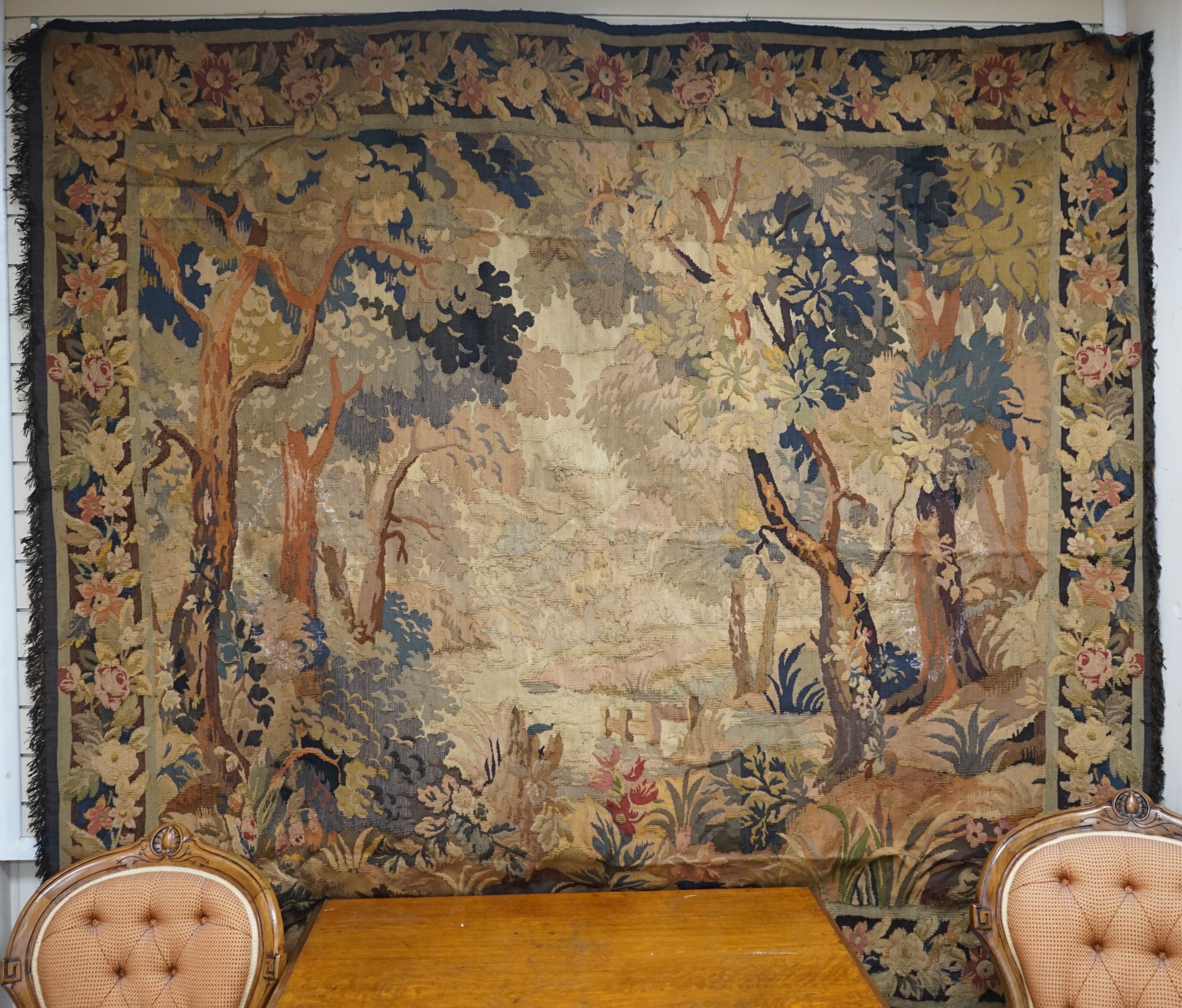 A late 19th / early 20th century French tapestry depicting a woodland scene with trees and flowers with a floral border woven in autumnal colours